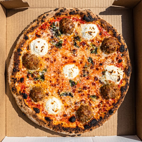 Neapolitan-style pizza topped with meatballs, dollops of ricotta, tomato sauce, melted cheese, and basil in a cardboard box.
