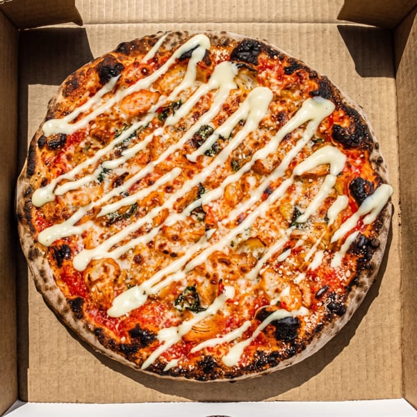 Freshly baked round pizza topped with melted cheese, vegetables, black olives, and white sauce drizzle in a cardboard box.