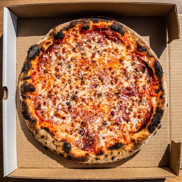 Large wood-fired pizza with charred crust, tomato sauce, melted cheese, pepperoni, and onions in an open cardboard box.